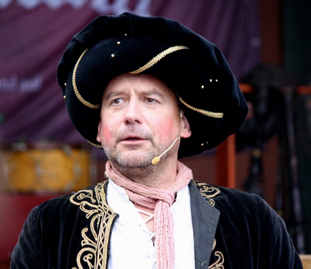 Birger Markuse in schwarzem Samtbarett mit Goldborte und rosa Schal, der Blick wandert zur Seite