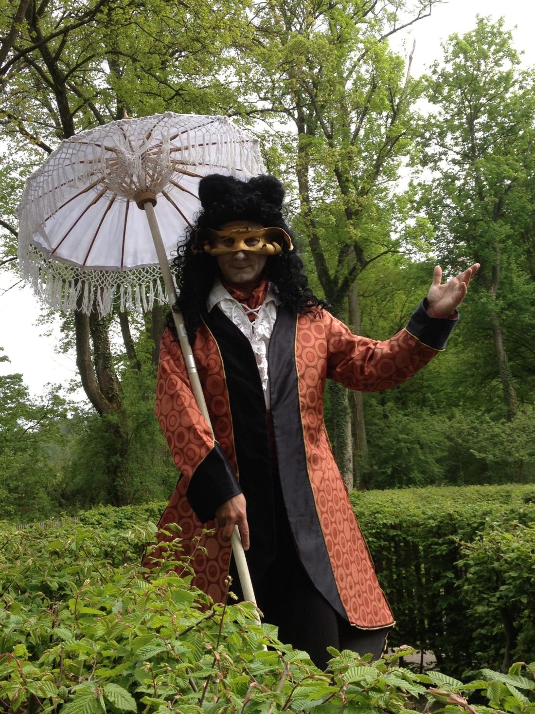 Stelzenfigur Monsieur de Parapluie mit Spitzenschirm, Maske und rotem Brokatmantel im Grünen