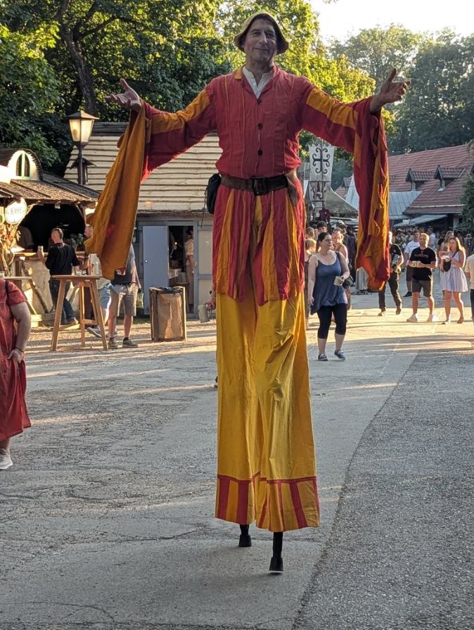 Stelzenfigur in Rot und Gelb mit ausgebreiteten Armen auf einem Mittelaltermarkt